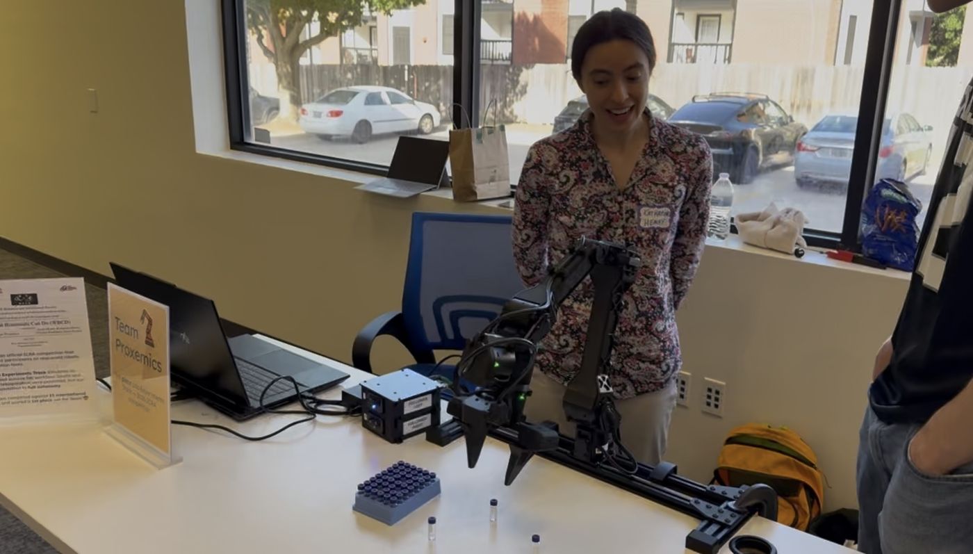 Katharine Henry demonstrating the robot arm to visitors at a table with a Team Proxemics sign.